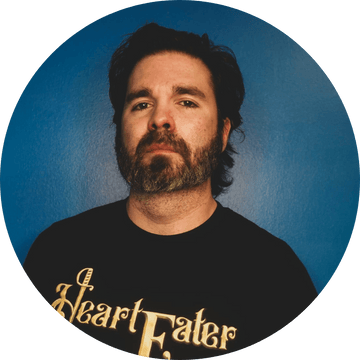 Confident man with a beard in a black Heart Eater t-shirt, posing against a blue background.