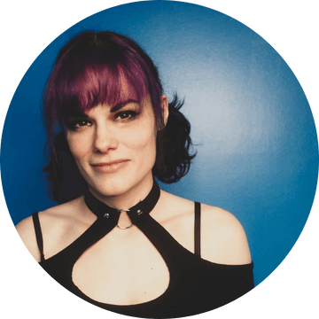 Portrait of a woman with purple hair and stylish attire against a blue background.